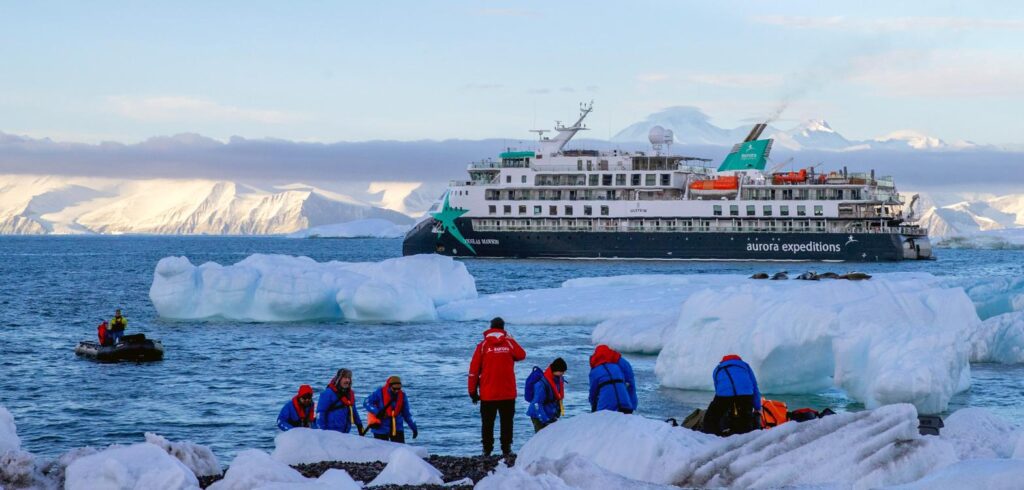 AE Expeditions’ Douglas Mawson lying off Cape Adare, Antarctica, with expeditioners in red parkas on the shore ice and a Zodiac running alongside