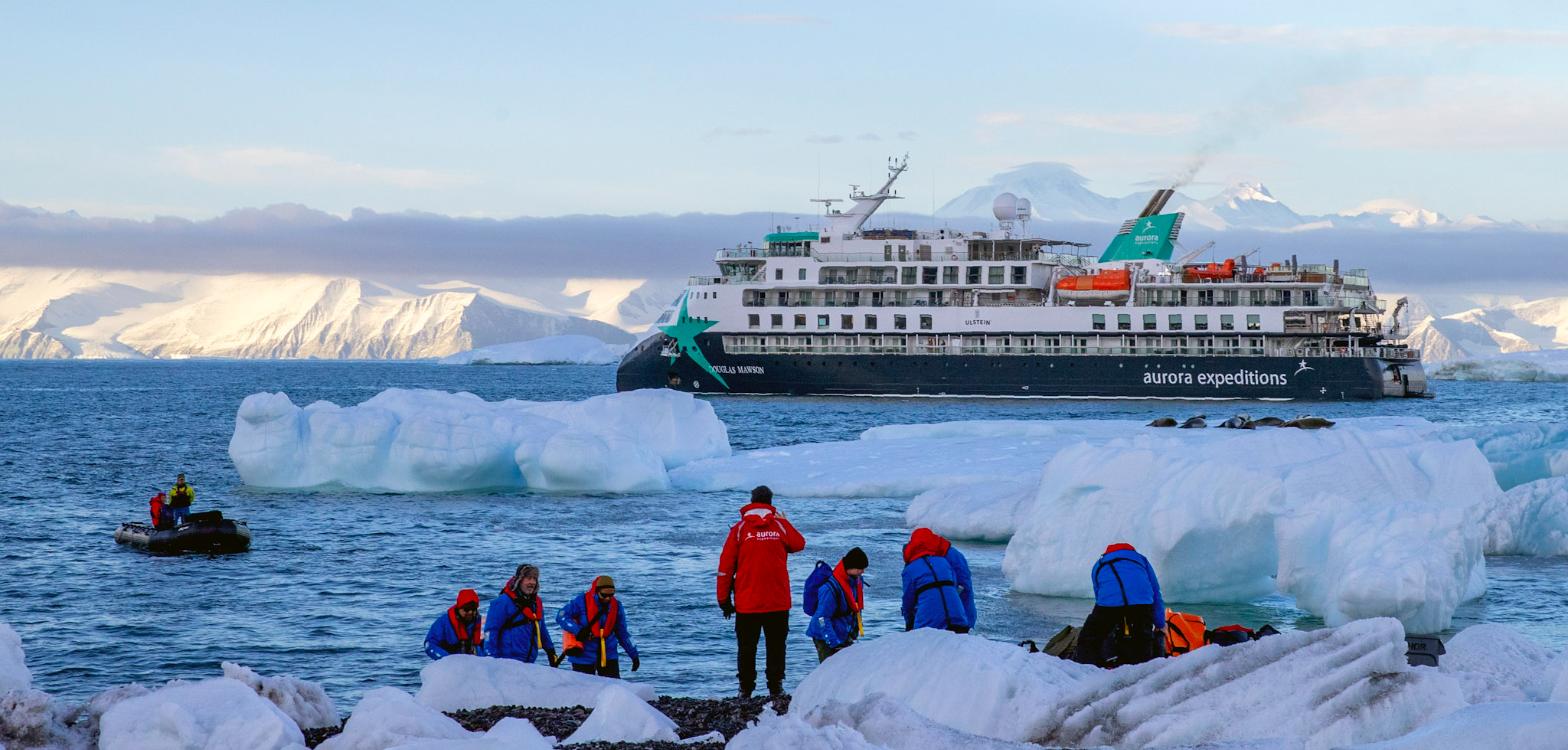 AE Expeditions’ Douglas Mawson lying off Cape Adare, Antarctica, with expeditioners in red parkas on the shore ice and a Zodiac running alongside