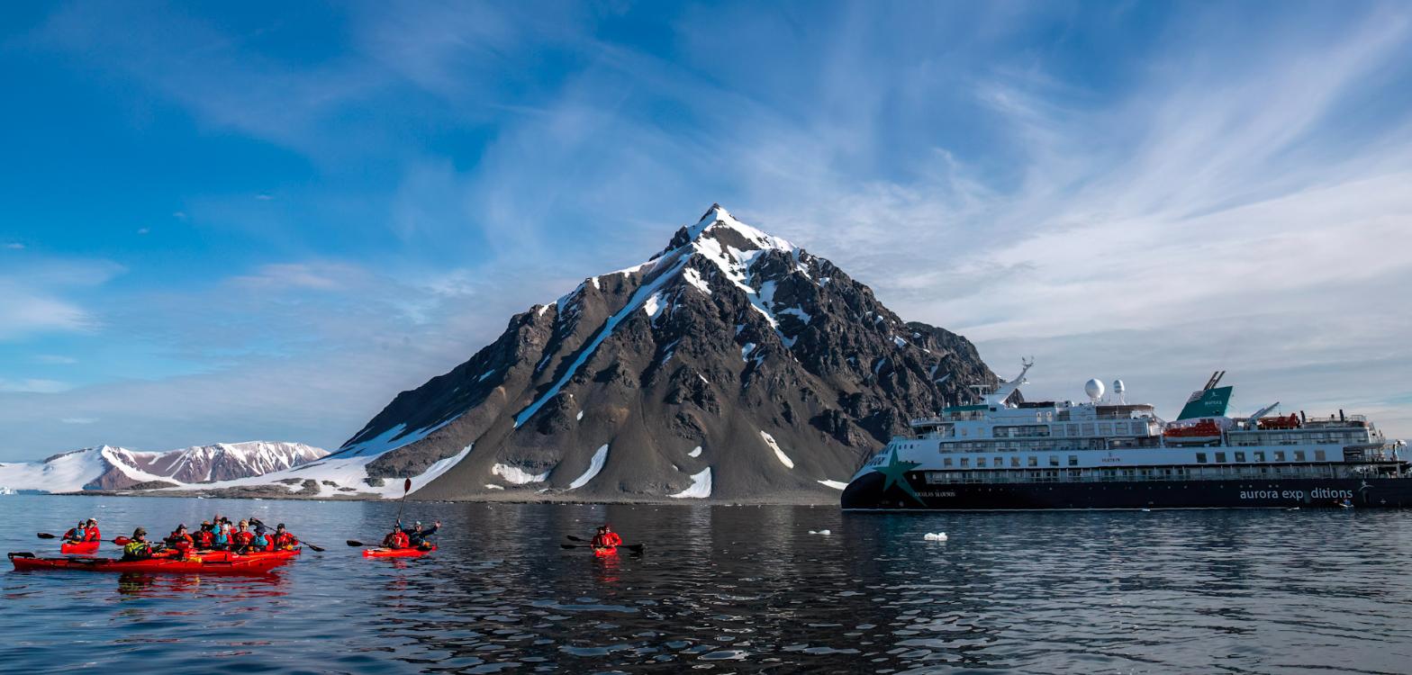 Guests kayaking in red sea kayaks beside AE Expeditions’ Douglas Mawson below a snow-streaked peak at Stonington Island, Antarctica
