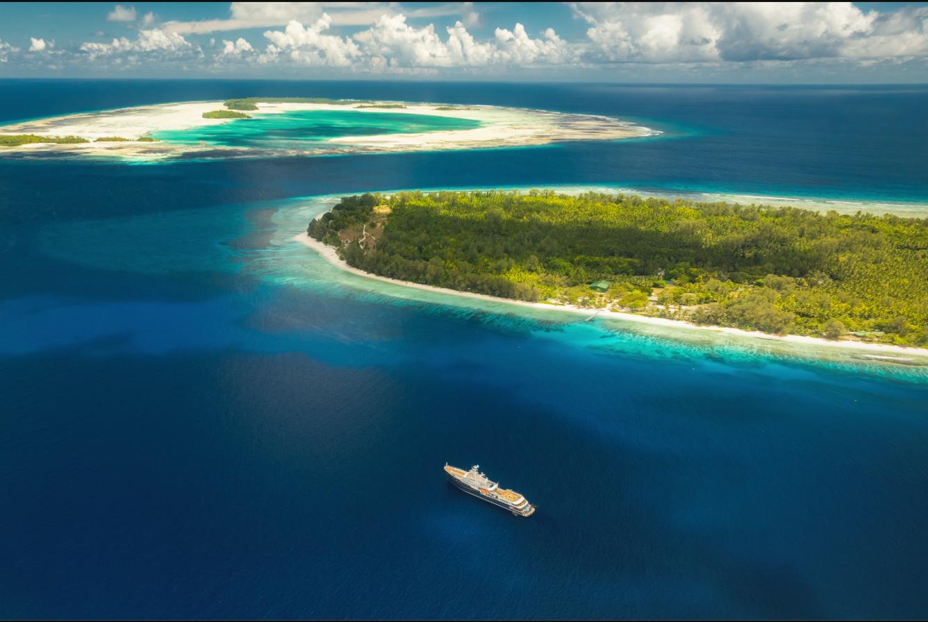 Aqua Lares anchored in deep blue water off a densely forested Seychelles island fringed by turquoise reef, with a low sand-rimmed atoll on the horizon beyond