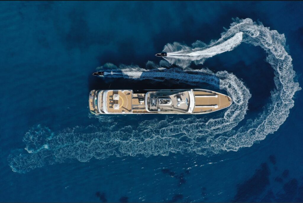 Overhead aerial view of the expedition yacht Aqua Lares under way in deep blue open water, her sun deck and helideck visible, with two tenders carving wakes alongside