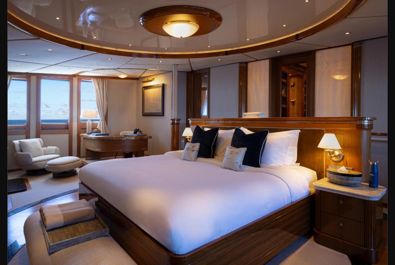An Owner's Suite aboard Aqua Lares, with a king bed under a curved wood-panelled ceiling, bedside reading lamps, and a writing desk and armchair beside windows looking out to sea
