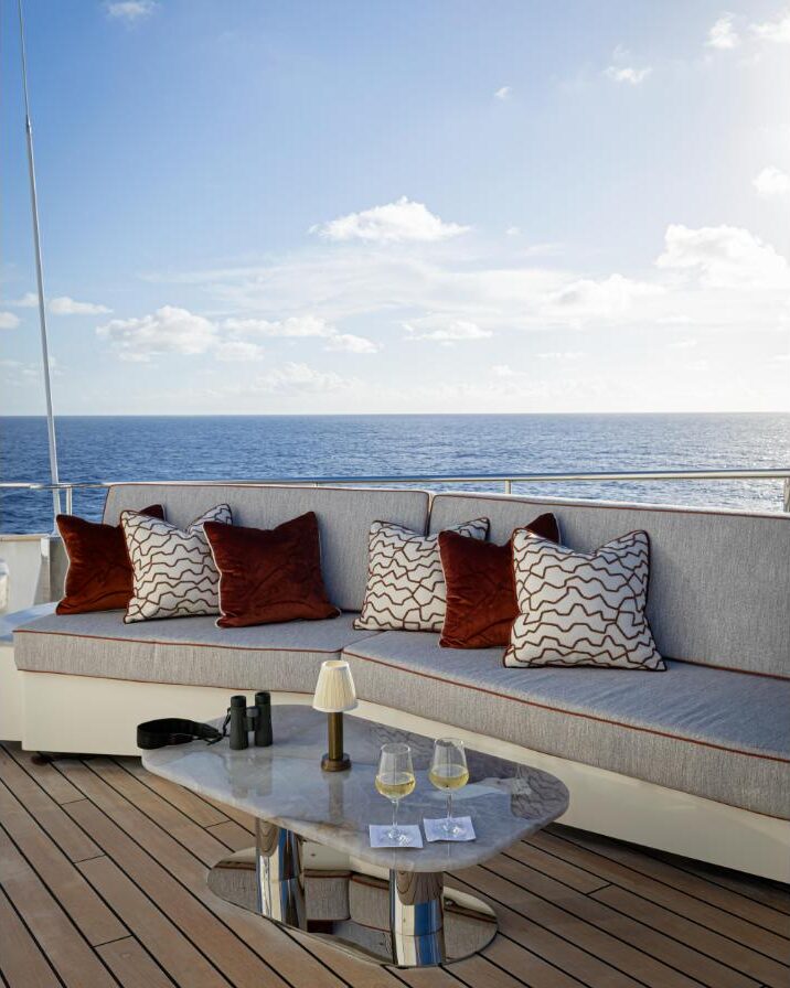 Curved grey sofa with rust and patterned cushions on the teak sun deck of Aqua Lares, with a marble table set with two glasses of white wine and binoculars, and open ocean beyond