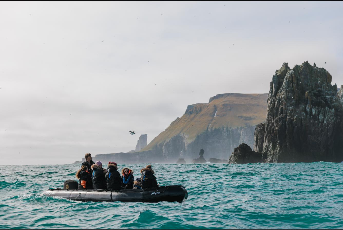 Guests in cold-weather gear aboard one of Aqua Lares' Zodiacs in open swell beneath steep basalt sea cliffs and rock stacks, with seabirds overhead