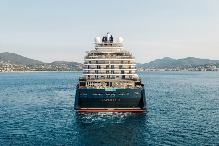 Explora Journeys Stretched Its Newest Ship by 19 Metres. Here’s What Went Into the Space EXPLORA III seen head-on at anchor in calm water, her navy hull and tiered white superstructure reflected in the sea, with a green coastline behind