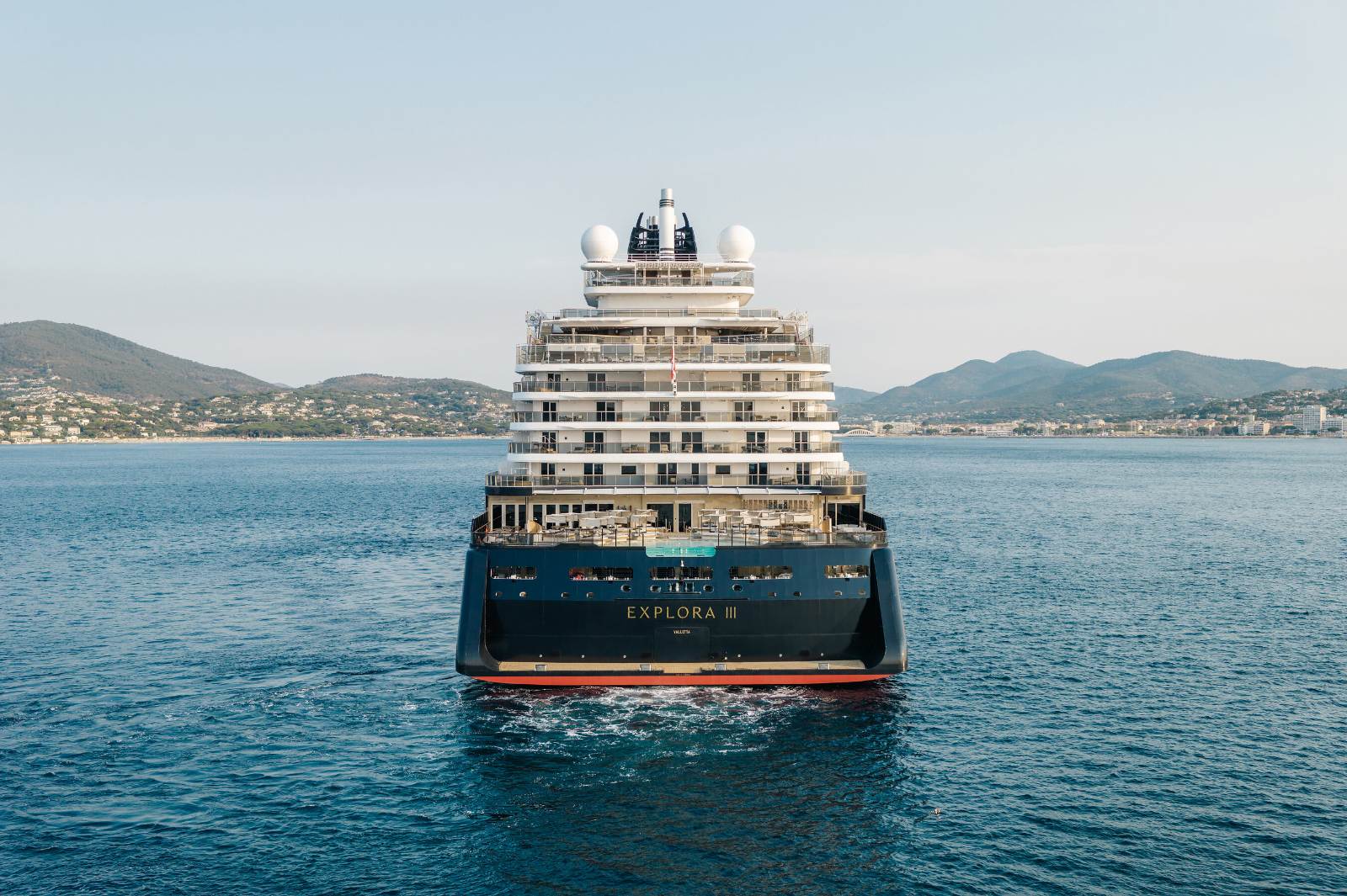 EXPLORA III seen head-on at anchor in calm water, her navy hull and tiered white superstructure reflected in the sea, with a green coastline behind