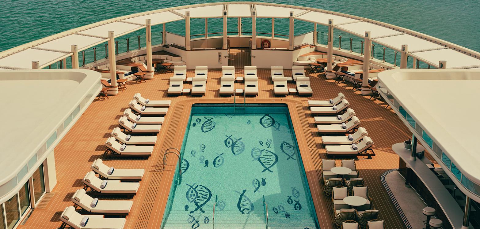 An overhead view of the pool deck on Four Seasons I: a rectangular pool with a blue fish mosaic, flanked by rows of white sun loungers on teak decking