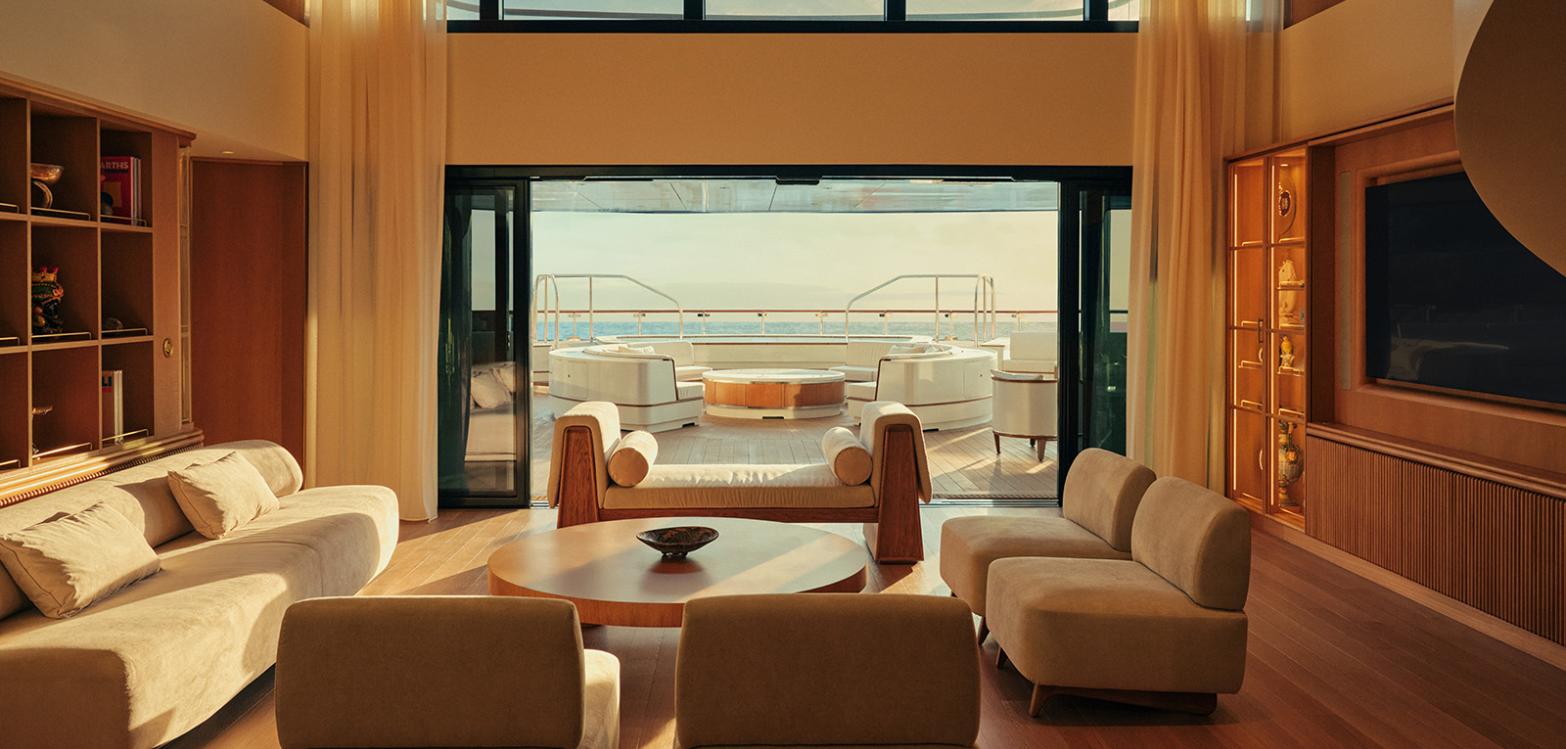 A double-height suite living room on Four Seasons I with pale suede sofas, a round timber coffee table, bookshelves and sliding glass doors opening onto a terrace with a daybed and the sea beyond