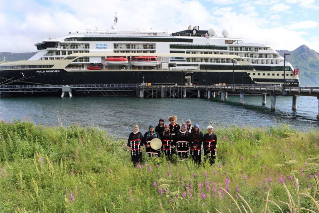 The first Old Harbor Alaska cruise call: MS Roald Amundsen alongside the pier at Old Harbor, with Alutiiq dancers in traditional regalia standing in the fireweed on the shore in front of the ship