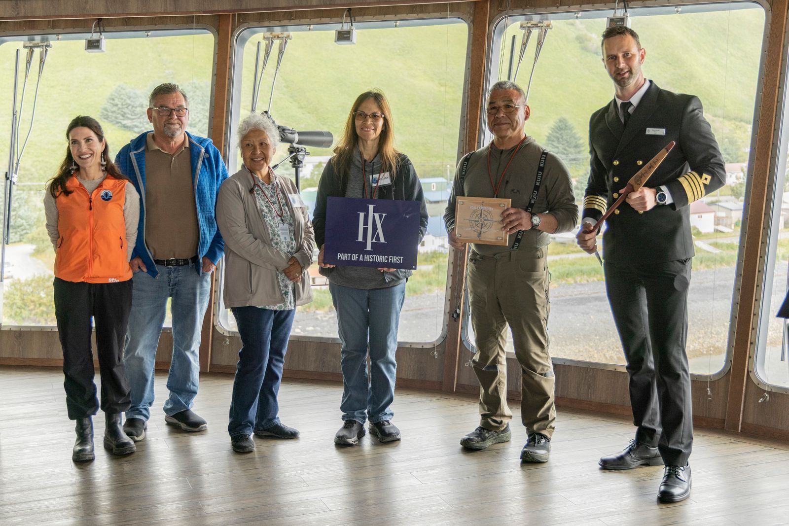 Six people, including an HX officer holding a ceremonial paddle, standing together indoors in front of windows looking out over Old Harbor village, with an HX banner between them
