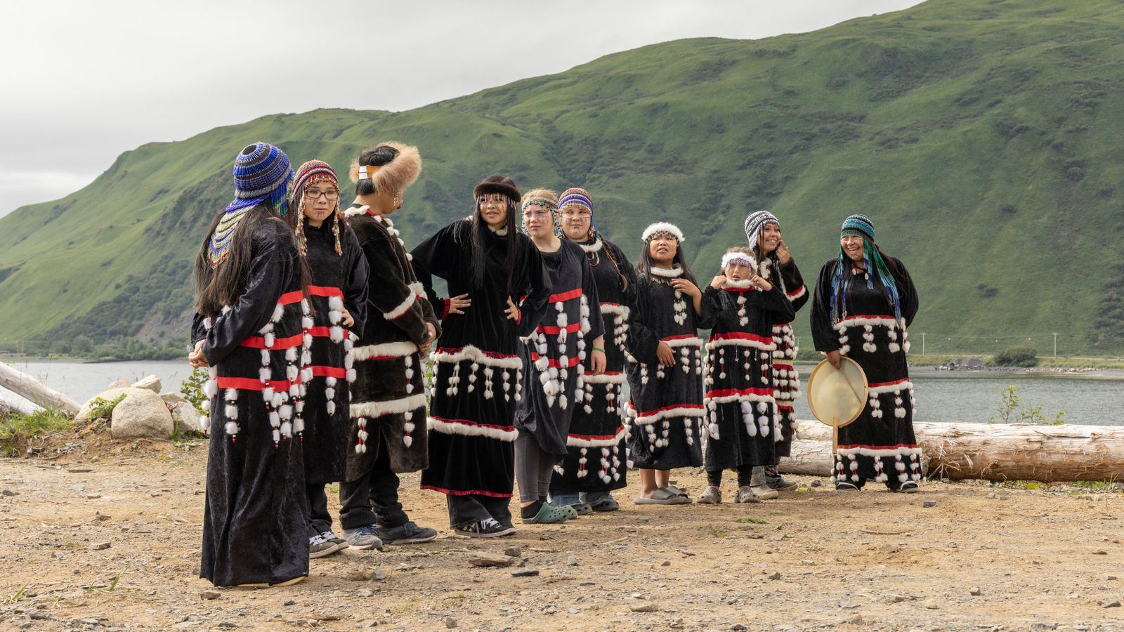 A group of Alutiiq dancers from Old Harbor on Kodiak Island in black regalia trimmed with white beadwork and fur, holding hand drums, with green hillsides behind them