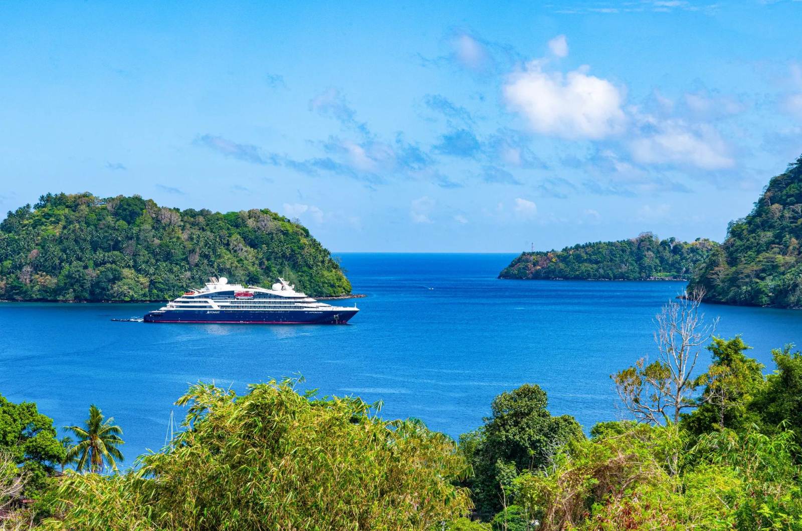 Ponant Indonesia itineraries call at Banda Neira: a Ponant ship at anchor in the bay, framed by jungle-covered headlands and bright blue water