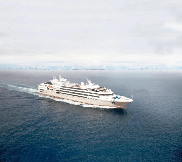Le Soléal under way on grey polar water, seen from above, her white superstructure and dark hull leaving a wake with an ice shelf on the horizon