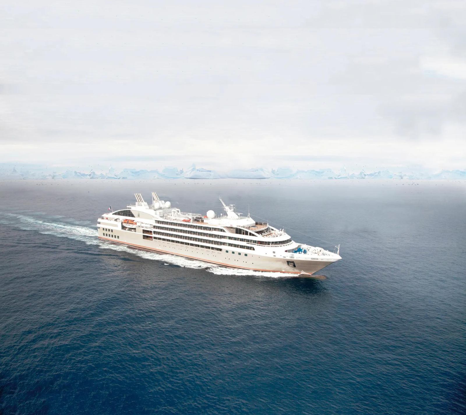 Le Soléal under way on grey polar water, seen from above, her white superstructure and dark hull leaving a wake with an ice shelf on the horizon
