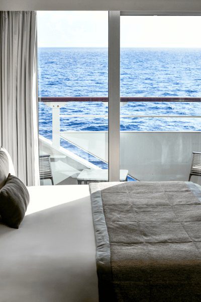 A Prestige Stateroom on Deck 5 of Le Soléal, with a grey-dressed bed, a bench seat and sliding glass doors onto a private balcony over open sea