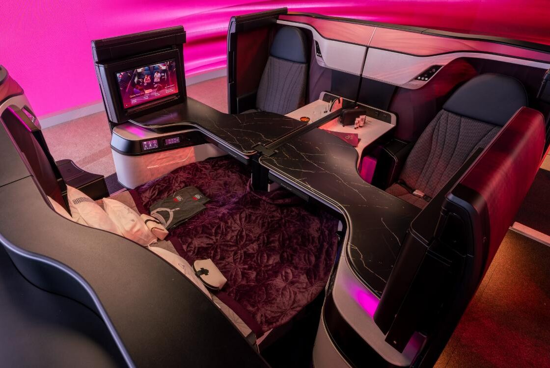 Two Qsuite Next Gen suites facing each other across a shared marble-effect dining table, with a bed made up alongside