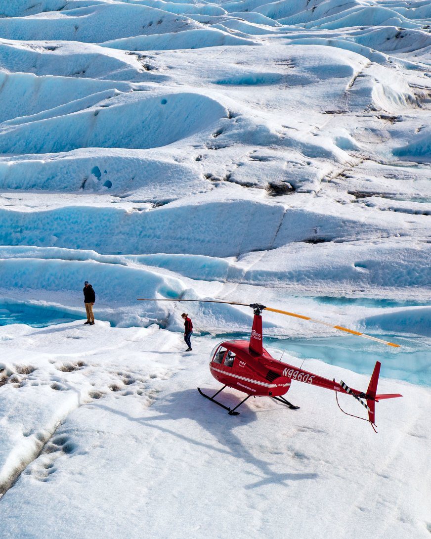 Alaska glacier helicopter landing, published by The Ritz-Carlton Yacht Collection to illustrate its Luminara Alaska cruise programme: two people on blue crevassed ice beside a red helicopter