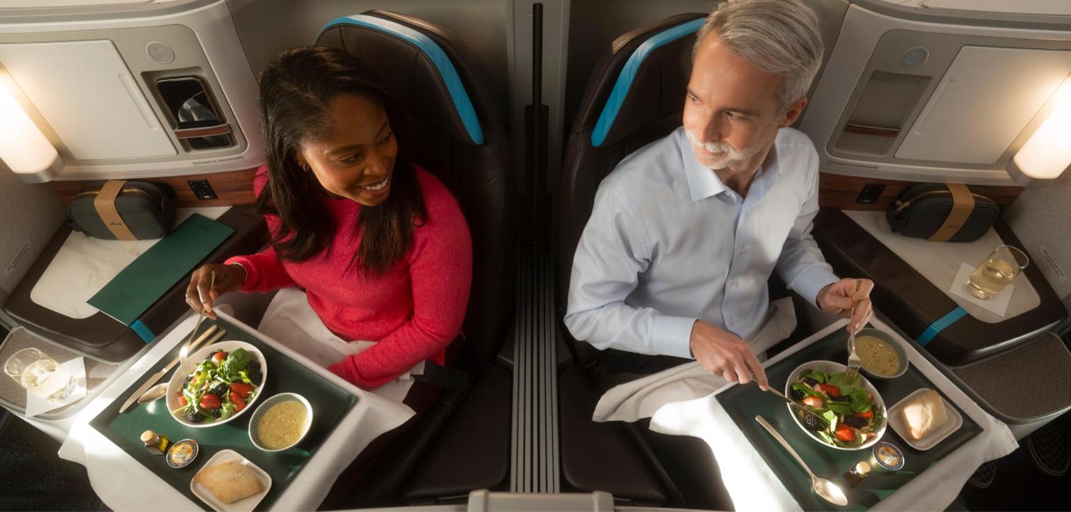 Two passengers eating salads at their seats in Alaska Airlines Boeing 787 Suites, with amenity kits and menus on the side consoles