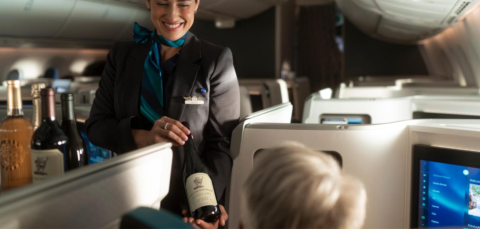 An Alaska Airlines flight attendant presenting a bottle of red wine to a passenger in the 787 Suites cabin, with more bottles on the counter beside her