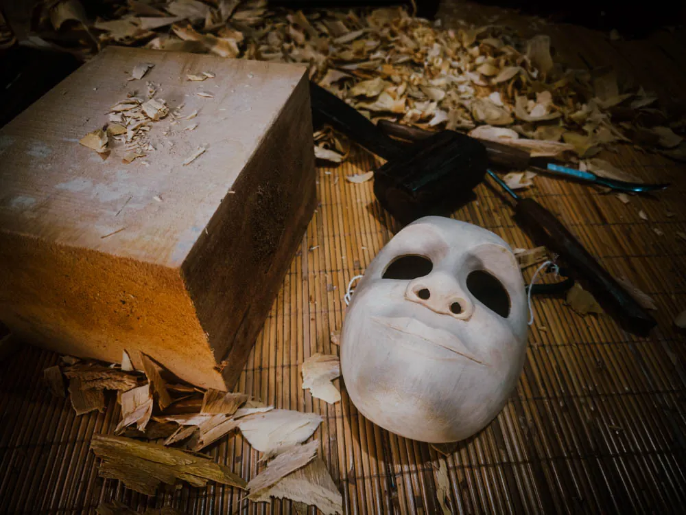 Unpainted carved wooden face mask next to an uncut block of wood and wood shavings