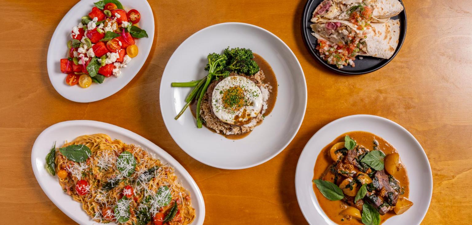 Overhead view of Hawaiian Airlines new chef-designed dishes, with Keaka Lee loco moco at the center, a watermelon salad, pasta, a braised beef dish and a wrap