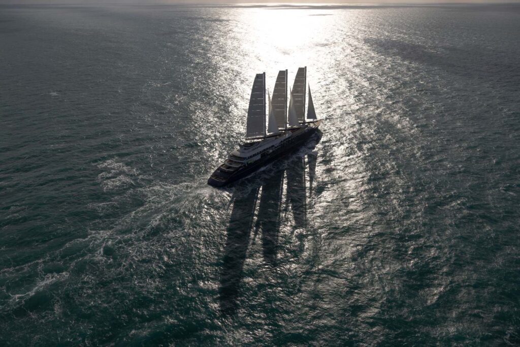 Aerial view of Orient Express Corinthian under its three rigid sails on open sea, with sunlight on the water